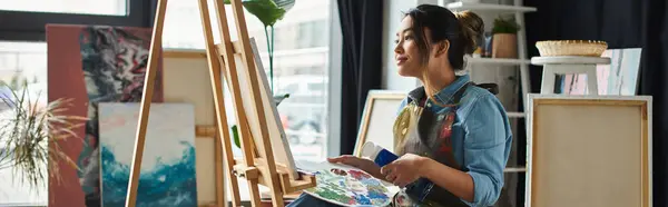 An Asian artist, wearing an apron, sits in her workshop, surrounded by canvases, lost in thought while holding a palette knife. — Stock Photo