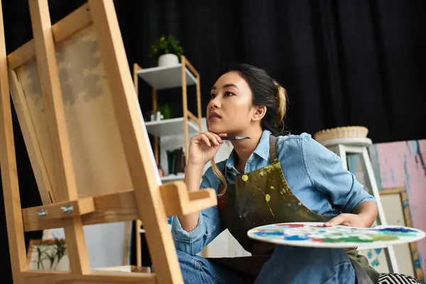 A young Asian woman, wearing an apron, sits in her art studio and thoughtfully contemplates her next brushstroke on a canvas. — Stock Photo