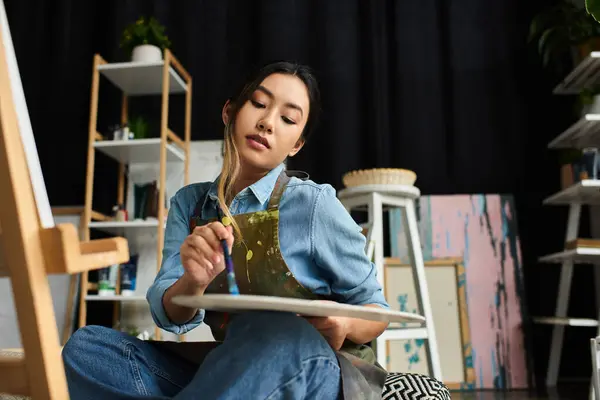 Азиатская художница в фартуке сидит в своей студии и смешивает краски на своей палитре. — стоковое фото