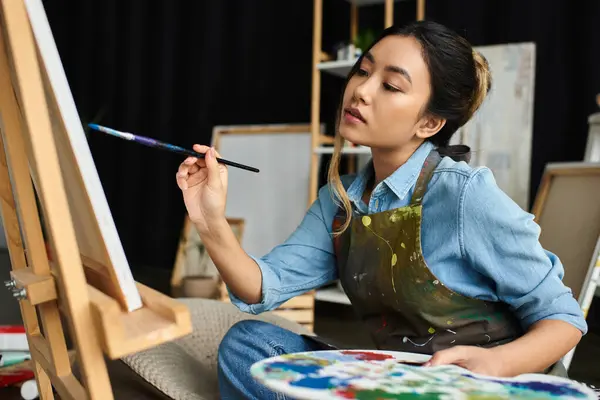 A young Asian woman in an apron is painting on a canvas in her workshop. — Stock Photo