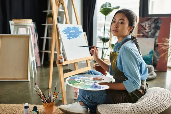 A young Asian woman in an apron sits in her art studio, painting on a canvas. — Stock Photo