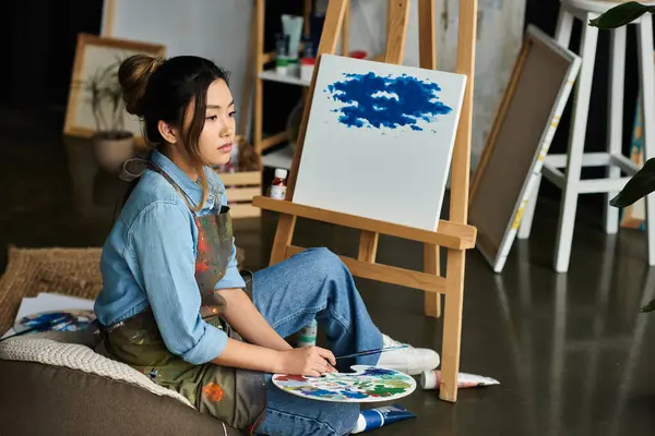 A young Asian woman, wearing an apron, sits in an art studio, contemplating her next stroke on her canvas. — Stock Photo