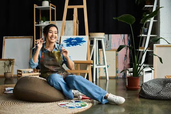 A young Asian artist takes a break from painting, sitting on a beanbag in her art-filled studio. — Stock Photo
