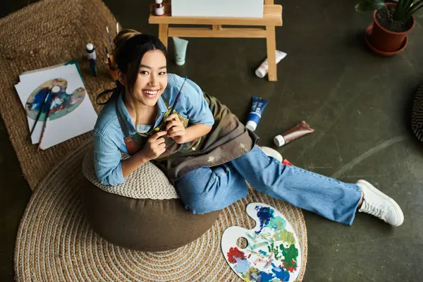 A young Asian woman, dressed in an apron, sits comfortably on a bean bag chair in her art studio, holding a paintbrush and smiling. — Stock Photo