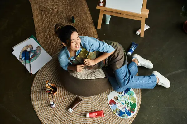 A young Asian woman in an apron relaxes on a beanbag chair in her art studio, holding a paintbrush. — Stock Photo