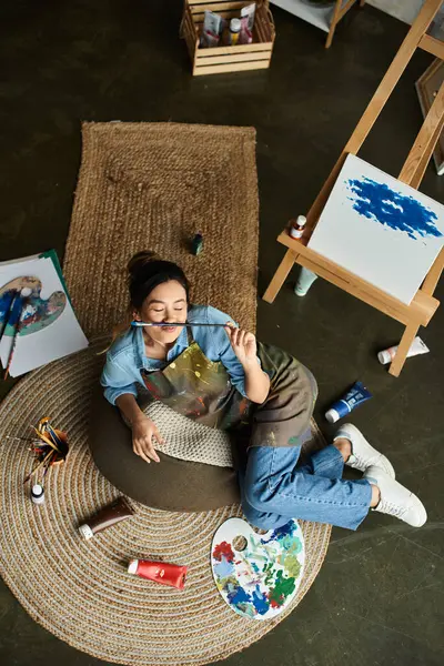 An Asian artist rests from painting, lounging on a beanbag with a paintbrush in her mouth, contemplating her work. — Stock Photo