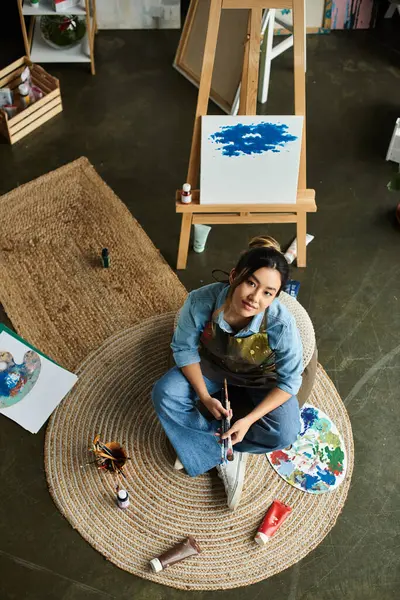 An Asian artist sits in her workshop, wearing an apron, and paints with brushes and paints. — Stock Photo