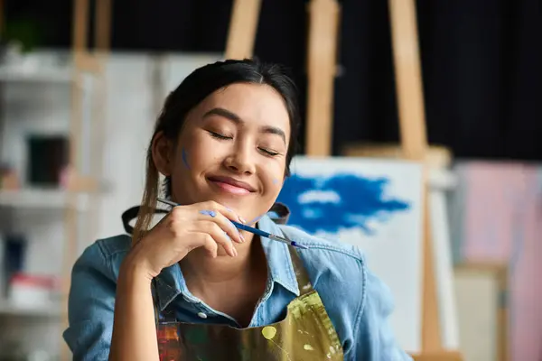 Une jeune artiste asiatique dans un tablier, perdue dans son travail, sourit en tenant un pinceau dans son atelier. — Photo de stock