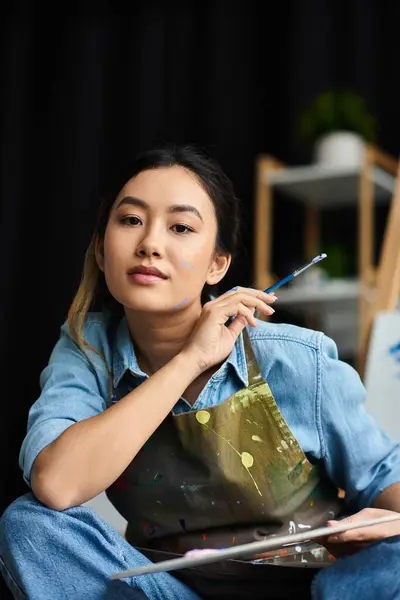 A young Asian woman in an apron sits in her art workshop, holding a paintbrush and contemplating her next stroke. — Stock Photo