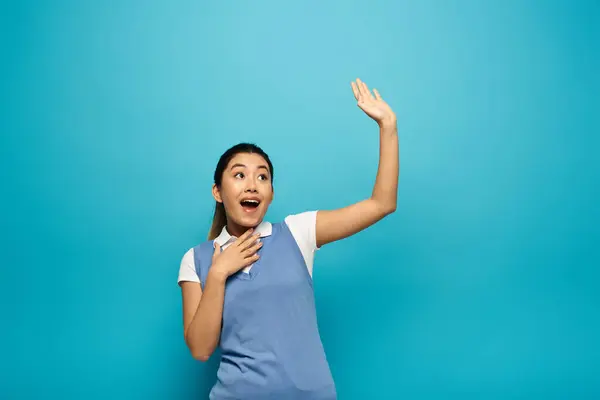 A surprised young Asian woman in smart casual attire stands against a blue background, mouth open and hand raised, looking up. — Stock Photo