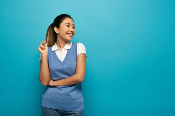 Uma jovem mulher asiática com uma camisa branca e colete azul sorri brilhantemente contra um fundo azul. — Fotografia de Stock