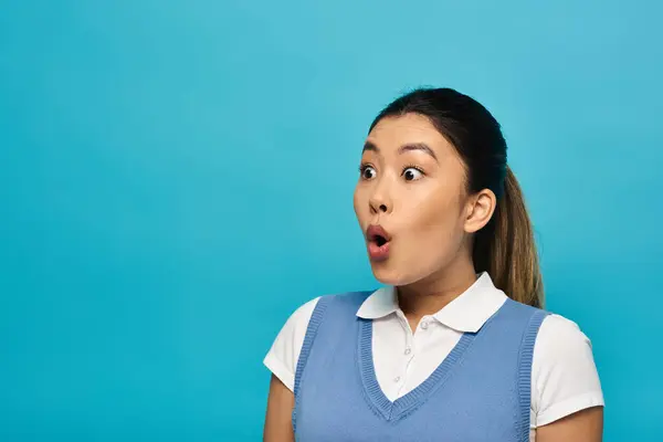A young Asian woman in smart casual attire stands against a blue background, looking surprised and with her mouth open. — Stock Photo