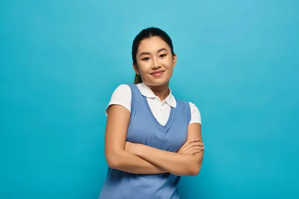 A confident young Asian woman in a white shirt and blue vest smiles at the camera with arms crossed against a blue backdrop. — Stock Photo