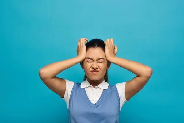 Uma jovem frustrada em trajes casuais inteligentes com cabelo escuro e um top azul segura a cabeça contra um fundo azul. — Fotografia de Stock