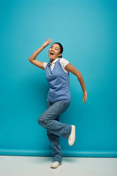 A young Asian woman wearing a blue vest and jeans stands against a blue background, smiling and laughing with her arm raised. — Stock Photo