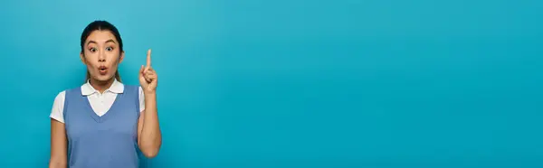 A young Asian woman in smart casual attire stands against a blue background, pointing upward with a surprised expression on her face. — Stock Photo