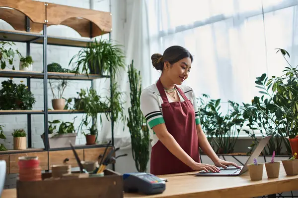 A smiling Asian woman wearing an apron works on laptop in her plant store, surrounded by greenery. — Stock Photo