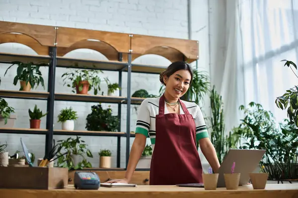A beautiful Asian woman, wearing an apron, stands behind the counter of her plant shop, surrounded by lush greenery. — Stock Photo