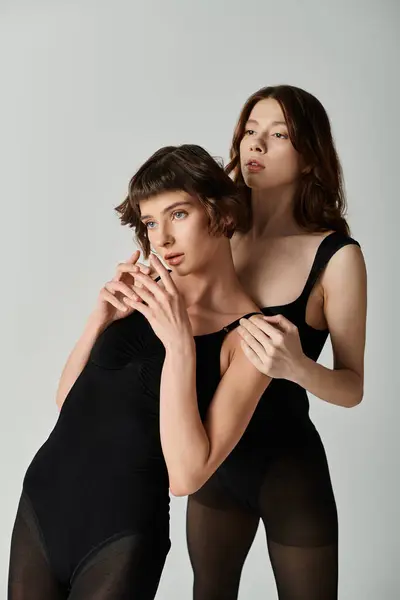 Two young women in black bodysuits pose against a grey backdrop, their bodies entwined in a loving embrace. — Stock Photo