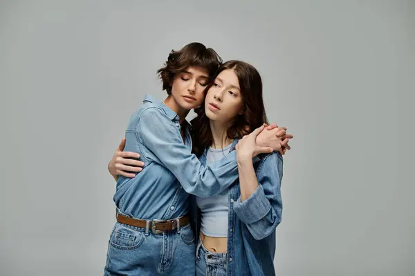 Two young women, dressed in denim, embrace each other on a grey background. — Foto stock