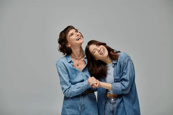 Two young women stand next to each other, arms wrapped around, showcasing denim attire on grey background. — Stock Photo