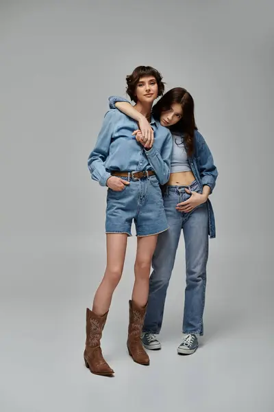 Two young women in denim outfits pose stylishly against a grey backdrop. — Foto stock