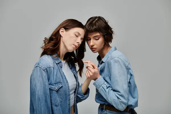 Two young women in denim attire pose intimately against a grey background, showcasing their love and style. — Stock Photo