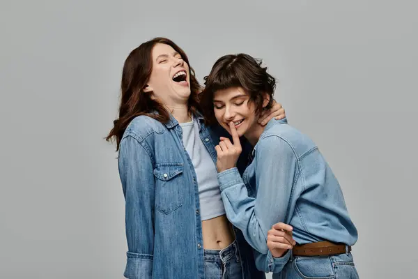 Two stylish young women in denim outfits pose against a gray background, laughing and enjoying each others company. — Stock Photo