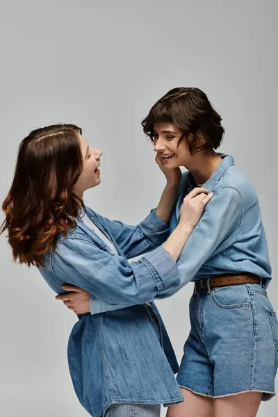 Two young women in denim attire pose against a grey backdrop, capturing a moment of playful intimacy and fashion. — Stock Photo