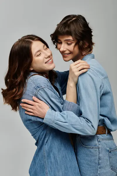 Two young women, dressed in denim, embrace and smile against a grey backdrop. — Stock Photo