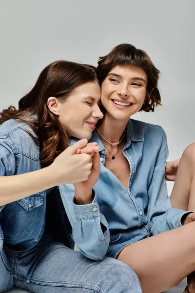 Two young women in denim attire pose affectionately against a grey backdrop. They are both smiling and look happy together. — Stock Photo