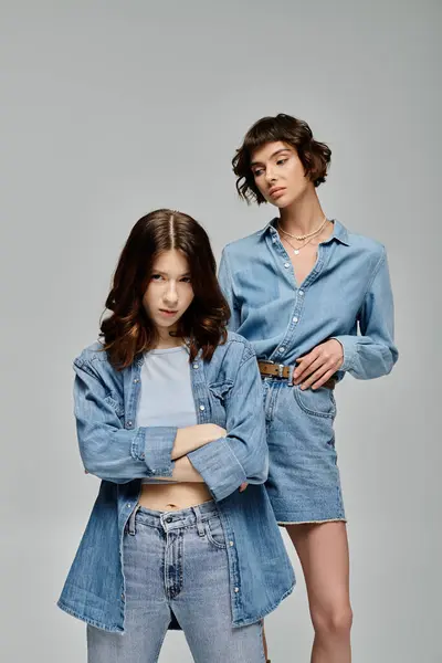Duas jovens mulheres em traje de ganga posar contra um pano de fundo cinza, apresentando um casal lésbico elegante e confiante. — Fotografia de Stock