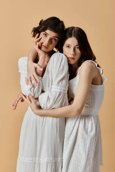 Two women in white dresses embrace against a beige background, showing love and affection. — Stock Photo