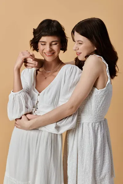 Дві молоді жінки в білих літніх сукнях обнімаються на бежевому фоні. Лесбійська пара — стокове фото