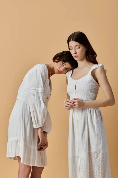 Two young women in white summer dresses stand close together on a beige background. — Stock Photo
