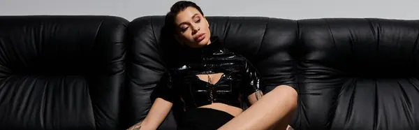 A young woman in sexy attire poses seductively on a black leather couch. — Stock Photo