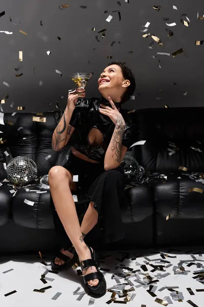 A woman in black sexy attire sits on a couch, celebrating with a drink, surrounded by falling confetti. — Stock Photo