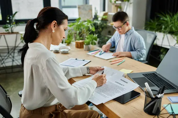 A mother works on a contract at her desk while her son with Down syndrome draws nearby. — Stock Photo
