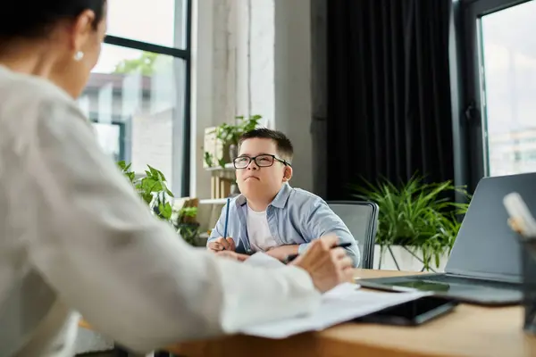 A mother works at her desk in an office while her son, who has Down syndrome, sits next to her. — Stock Photo