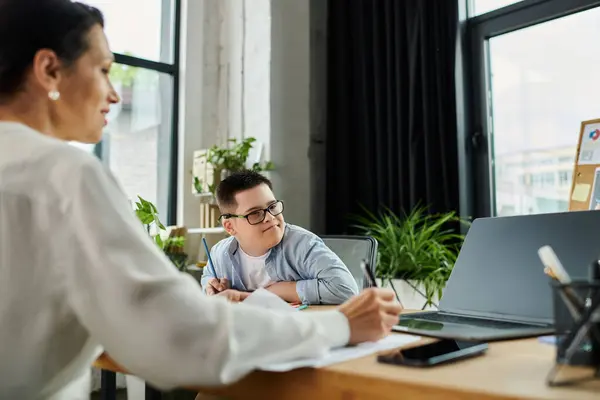 Une mère travaille sur son ordinateur portable pendant que son fils trisomique est assis à côté d'elle à un bureau. — Photo de stock