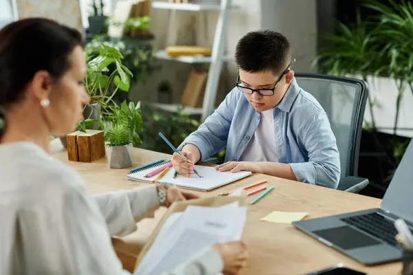 Eine Mutter arbeitet an ihrem Schreibtisch, während ihr Sohn mit Down-Syndrom am selben Tisch zeichnet und eine Familie zeigt, die Vielfalt und Inklusion umarmt. — Stockfoto