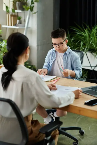 Une mère travaille à son bureau tandis que son fils trisomique est assis à côté d'elle, apprenant et coloriant. — Photo de stock