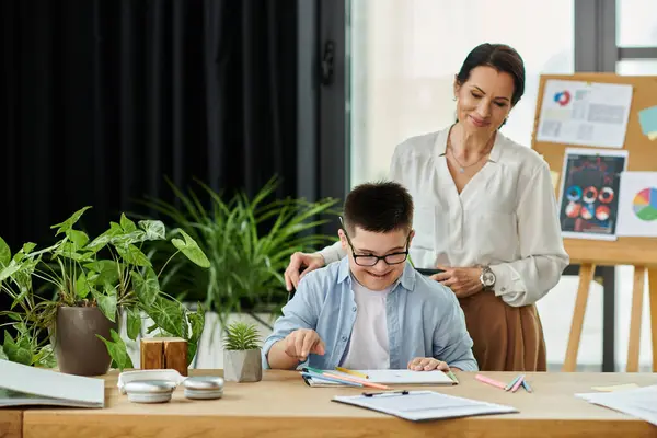 Una madre está detrás de su hijo con Síndrome de Down mientras él trabaja en un escritorio en un entorno de oficina. - foto de stock