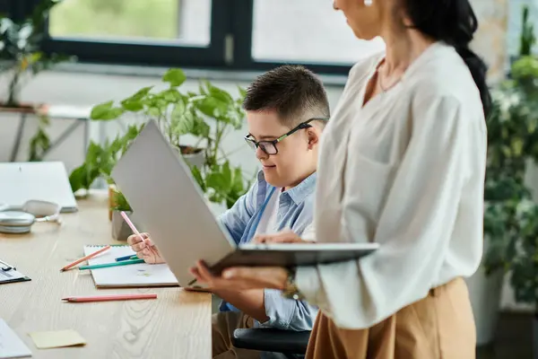 Une mère travaille à un ordinateur portable dans un bureau tandis que son fils avec des couleurs trisomiques à proximité. — Photo de stock
