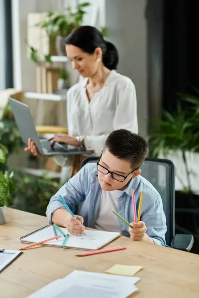 Un jeune garçon trisomique dessine à une table tandis que sa mère travaille sur un ordinateur portable en arrière-plan. — Photo de stock