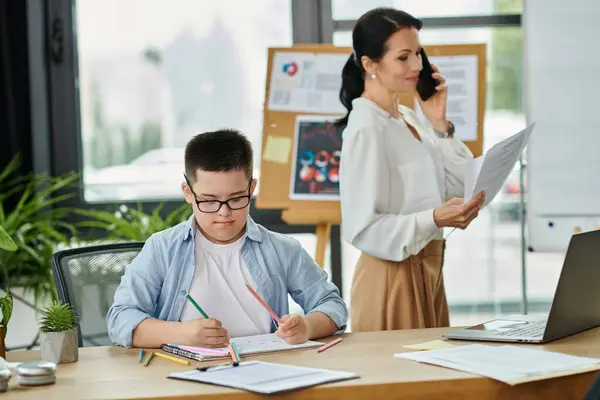 A mother works on the phone and paperwork while her son with Down syndrome sits at a desk drawing in an office. — Stock Photo
