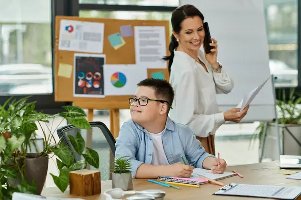Une mère travaille dans un bureau, concentrée sur un appel téléphonique, tandis que son fils atteint du syndrome de Down est assis à un bureau à colorier. — Photo de stock
