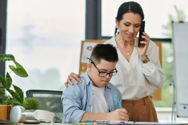 Mother works in office while son with Down syndrome studies nearby, emphasizing inclusivity and support in the workplace. — Stock Photo