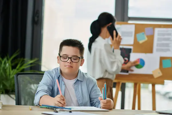 Un jeune garçon trisomique est assis à un bureau, dessinant avec des crayons de couleur, tandis que sa mère travaille dans le bureau derrière lui. — Photo de stock