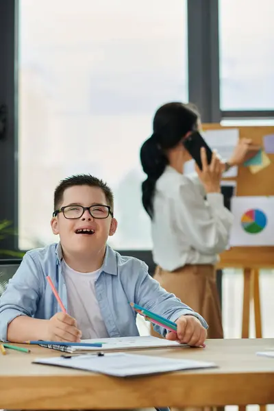 Un jeune garçon trisomique sourit alors qu'il colore à un bureau, tandis que sa mère travaille en arrière-plan. — Photo de stock